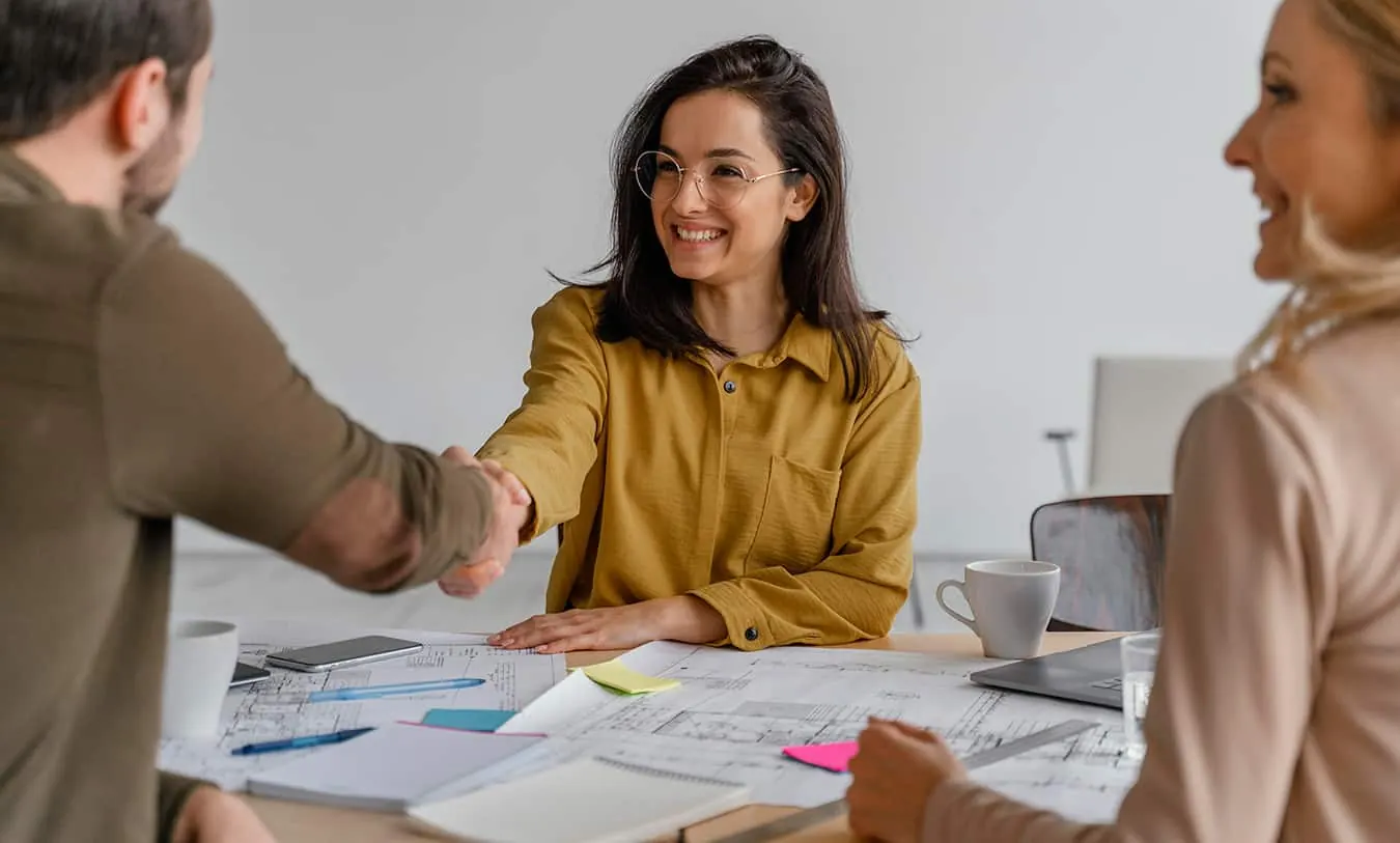 A smiling woman shakes hands across a table at a business meeting.
