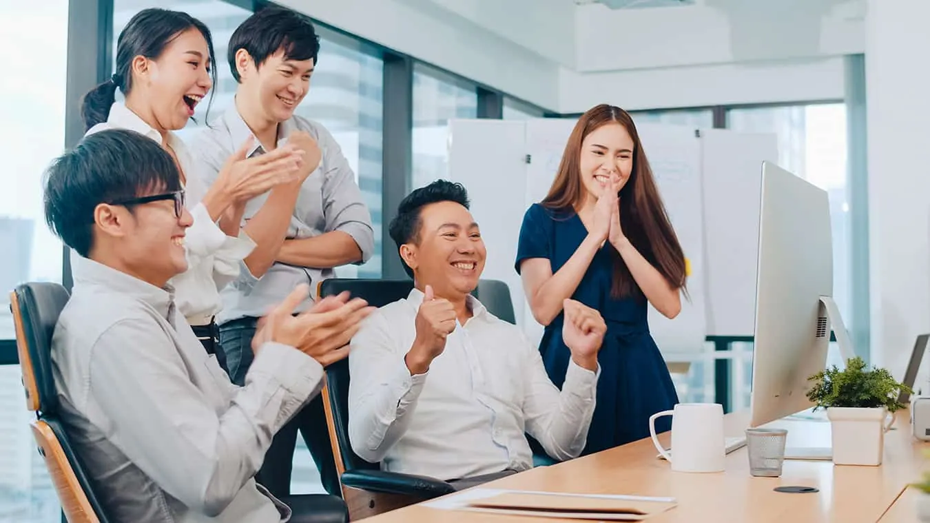 A young business team claps and celebrates around an office desk.