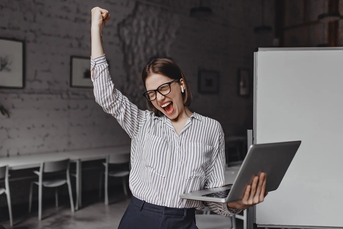 A businesswoman in glasses cheers with one arm raised holding a laptop.