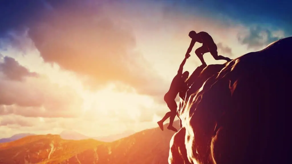 One climber pulls another up a rocky mountain peak at sunset.