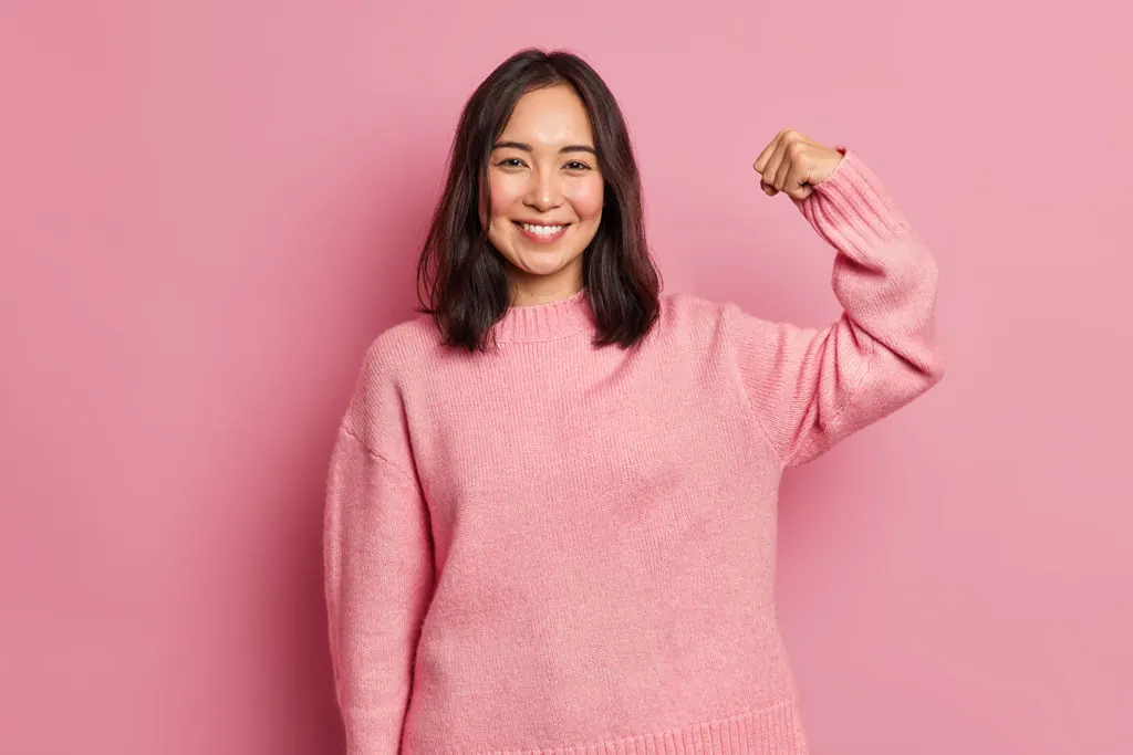 A smiling woman in a pink sweater flexes her arm on a pink background.