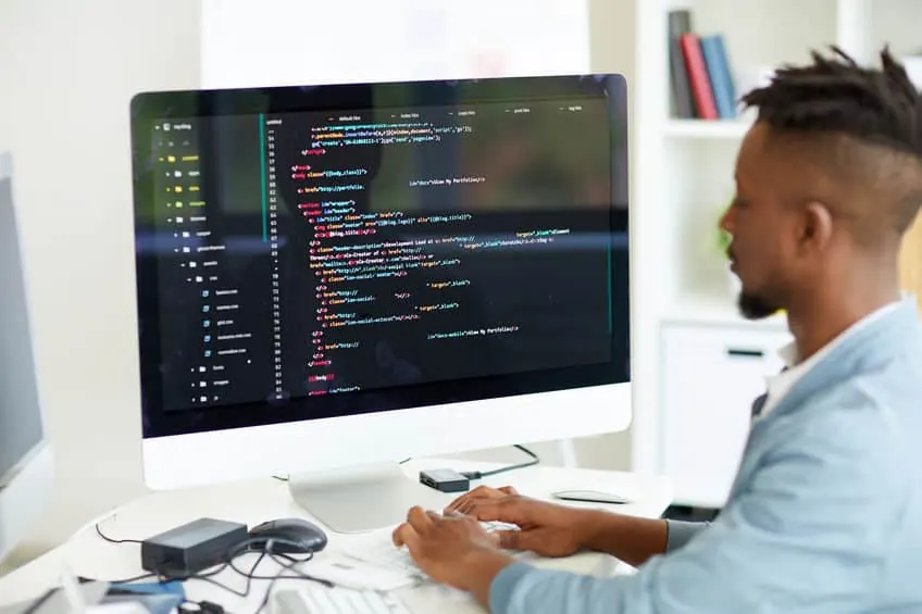 A developer writes code on a large desktop monitor at a bright desk.