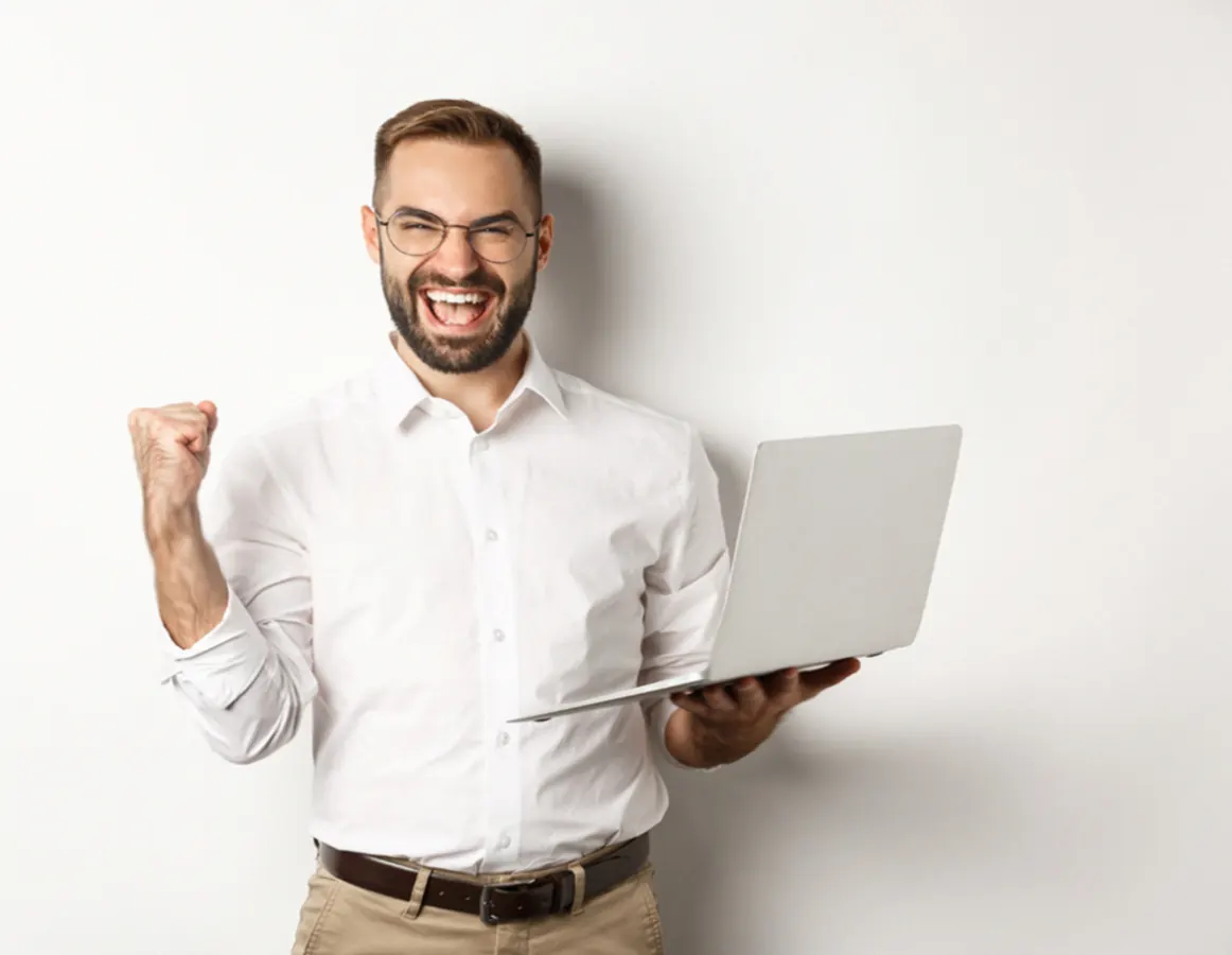 An excited man in a white shirt holds a laptop and cheers.