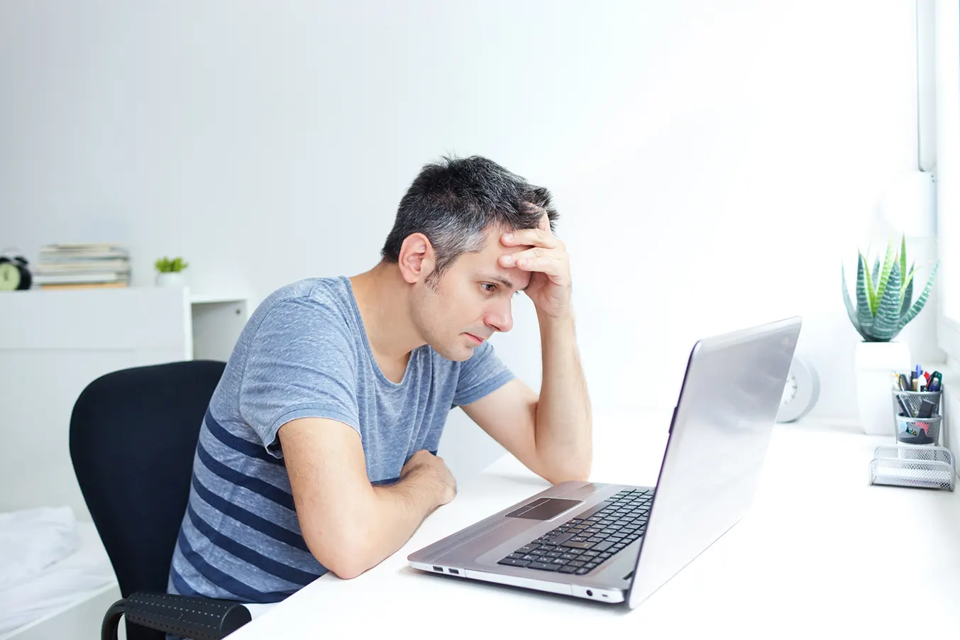A frustrated man holds his head while looking at his laptop.