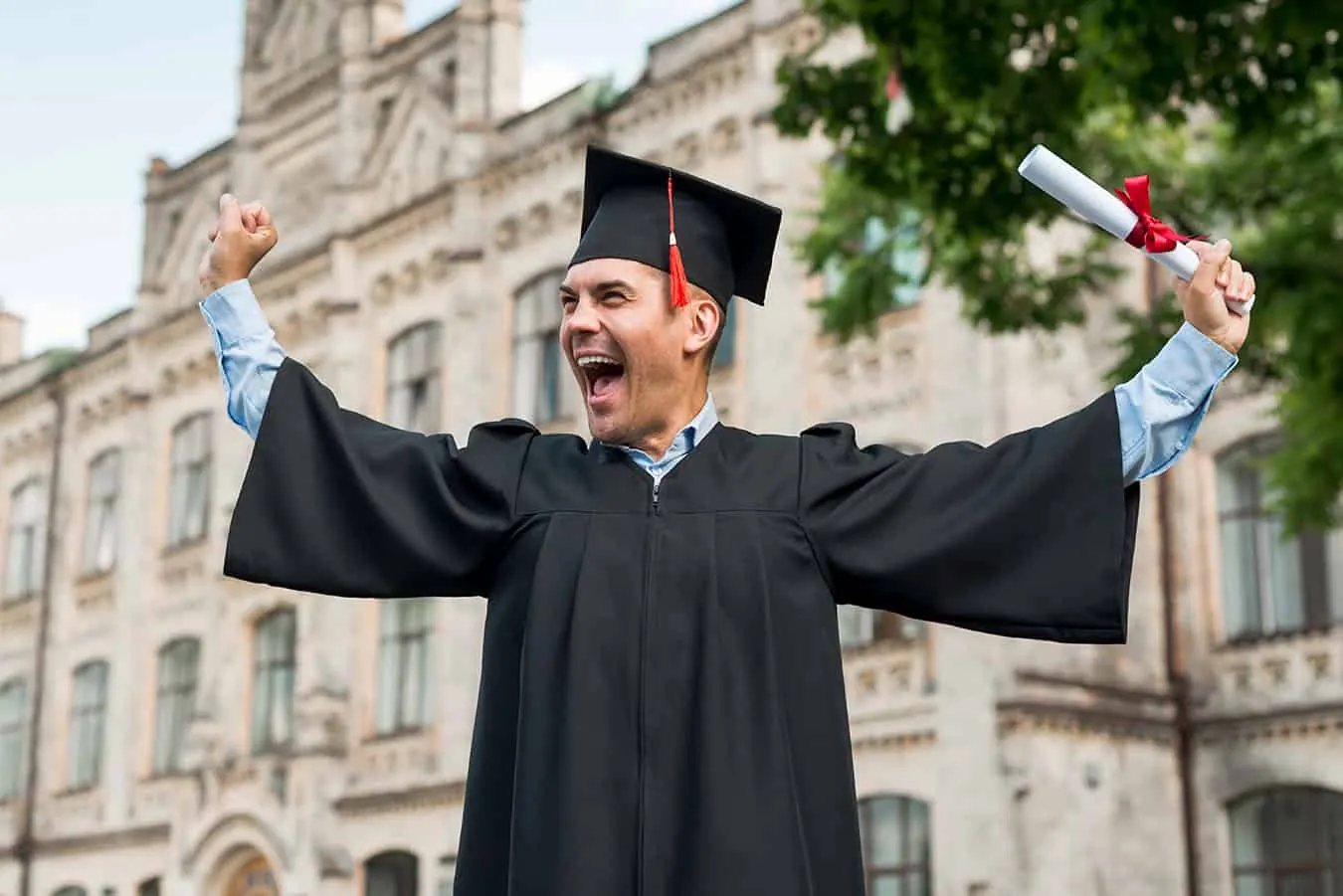 A graduate in a cap and gown cheers holding a diploma.