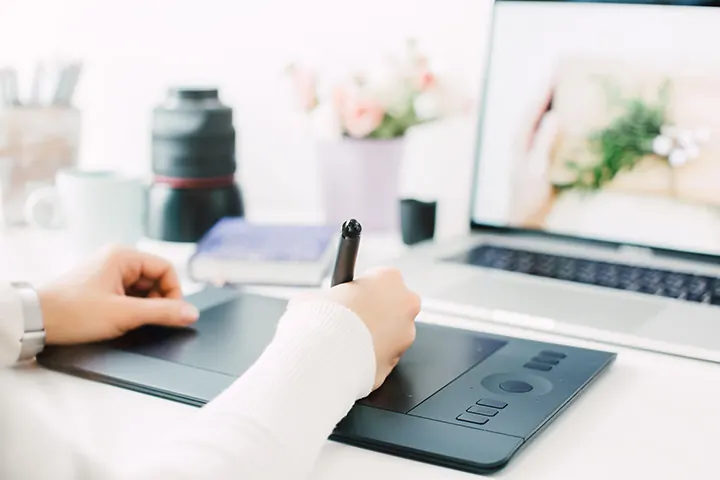 A hand draws with a stylus on a graphics tablet near a laptop.