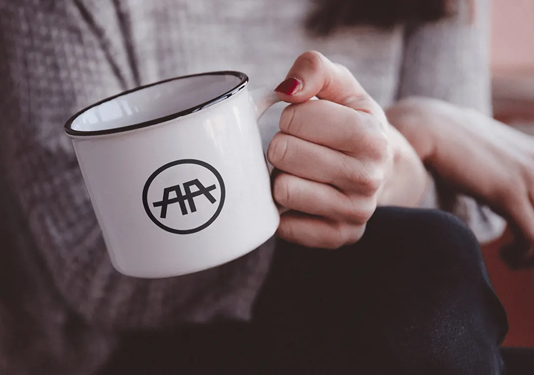 A hand holds a white enamel mug printed with the AA logo.