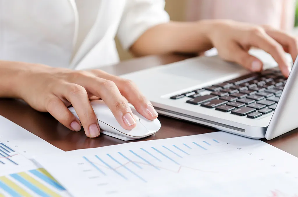 Hands using a laptop keyboard and a white mouse on a desk.
