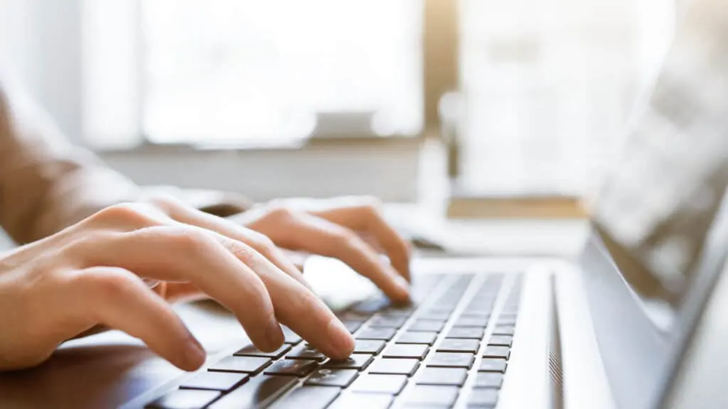 Close-up of hands typing on a silver laptop keyboard near a window.