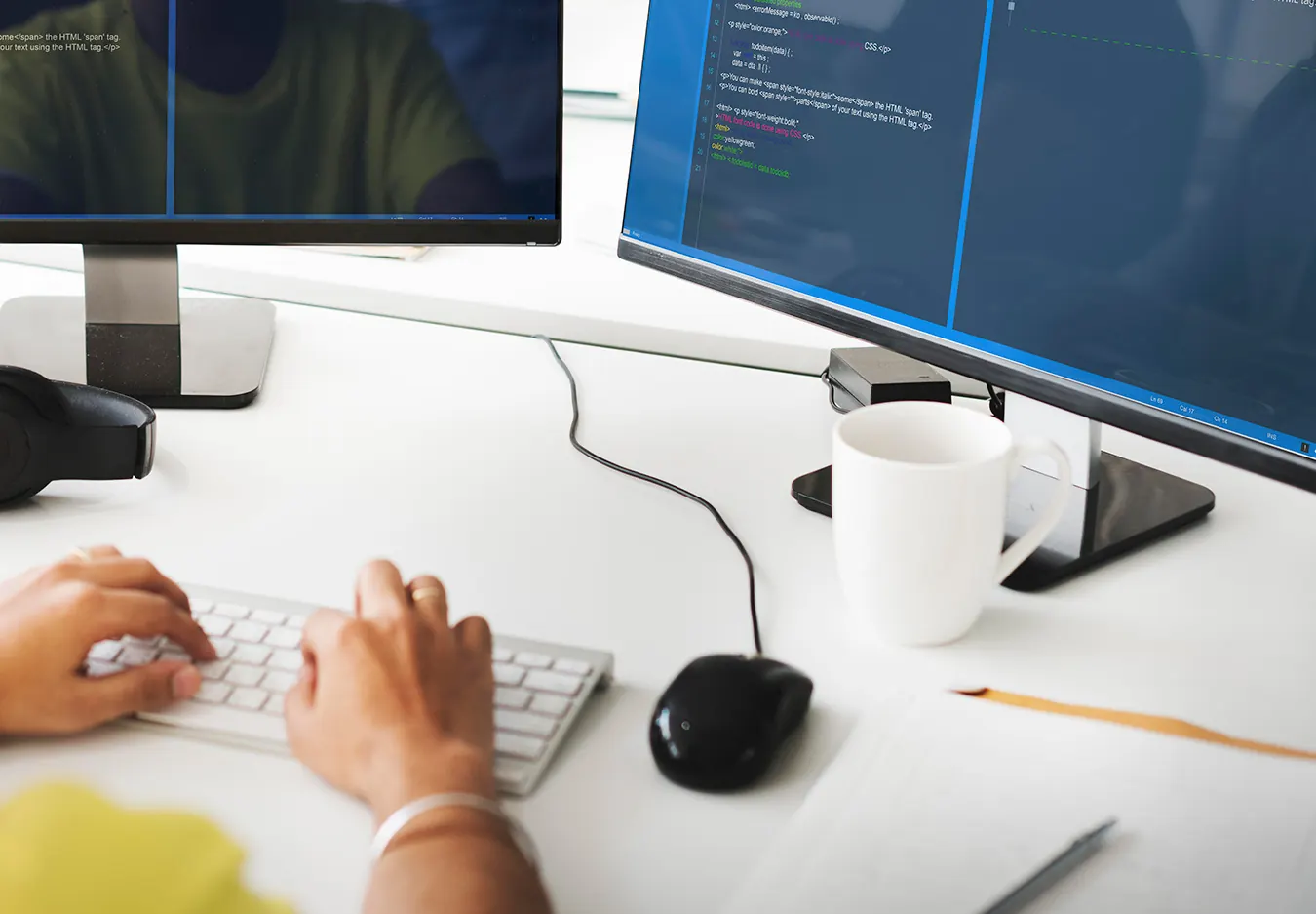 Hands type on a white keyboard at a desk with two monitors.