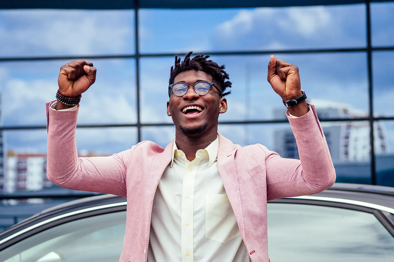 A smiling man in a pink blazer raises both fists in celebration.