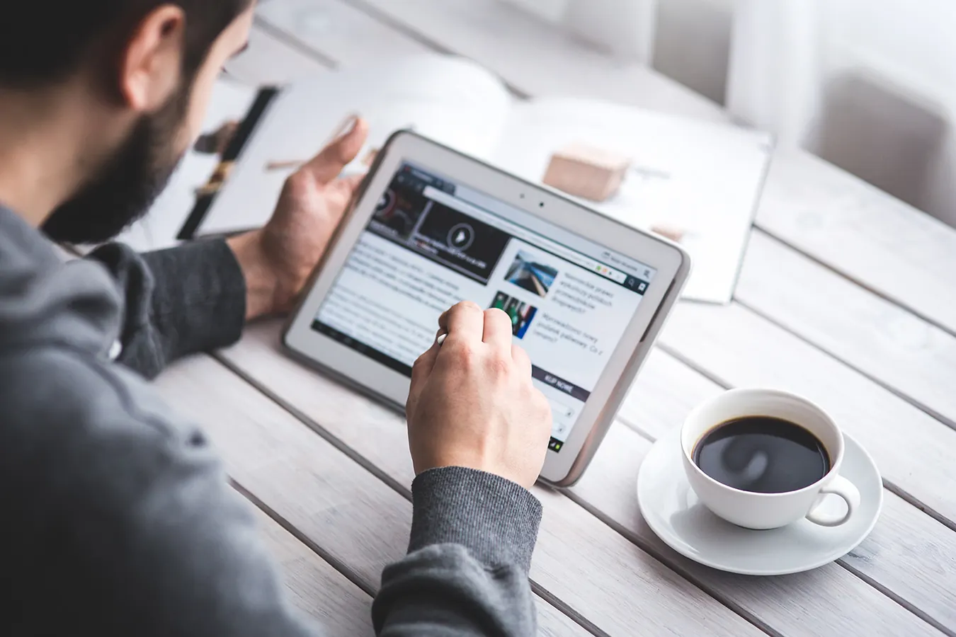 A man reads news on a tablet at a table with a coffee cup.