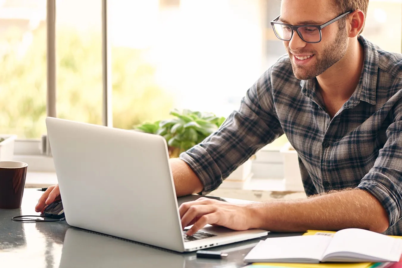 A smiling man in glasses works on a laptop at a bright desk.