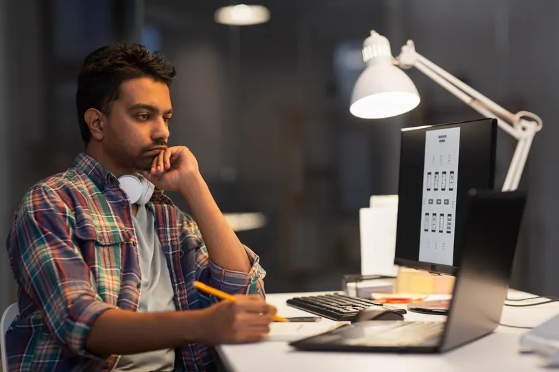 A man in a plaid shirt thinks while writing beside a laptop.