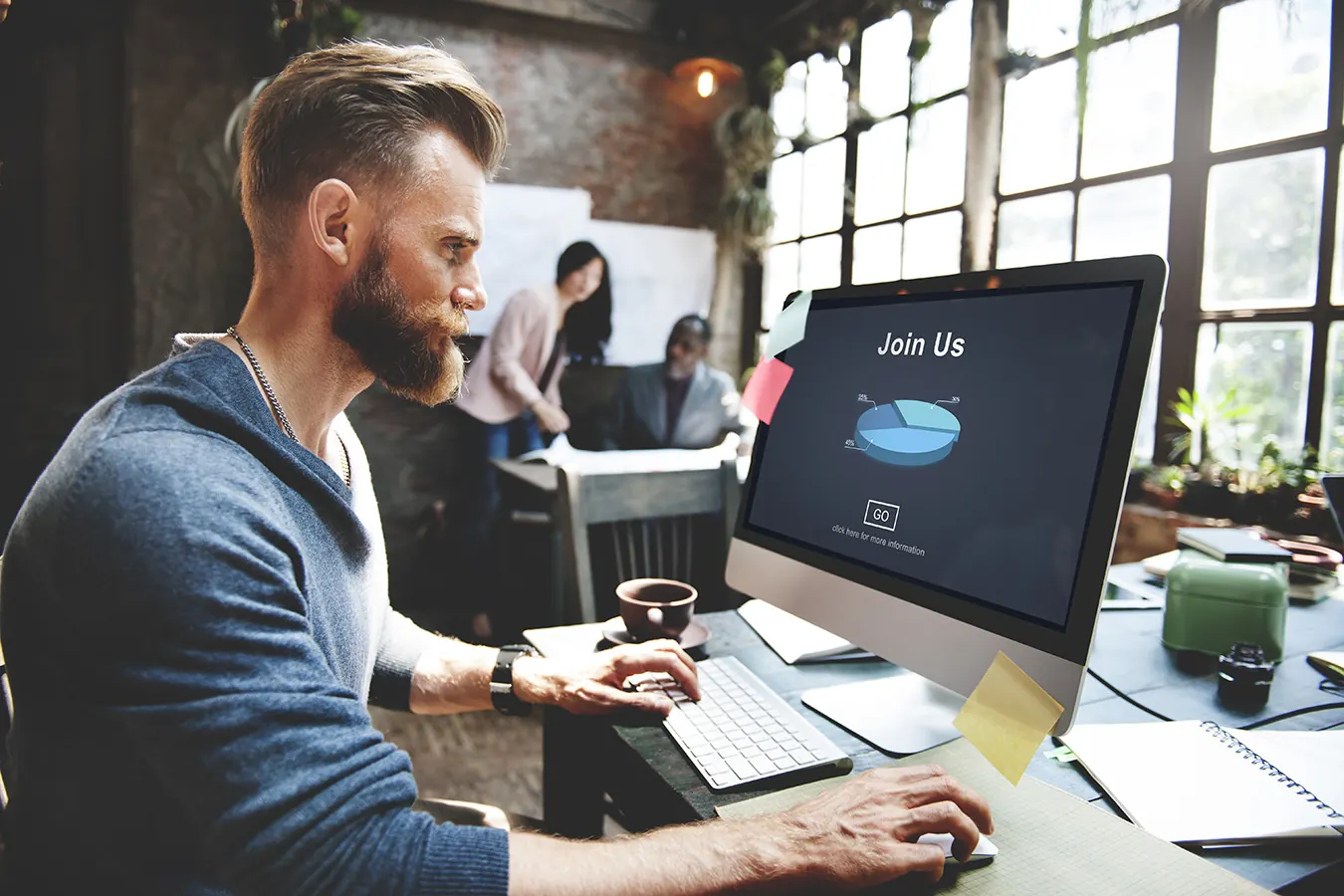 A bearded man types at a desktop computer in a loft office with windows.