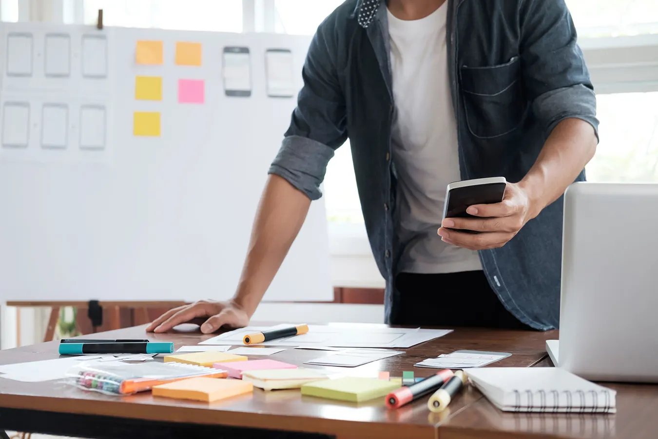 A man checks his phone while standing at a desk covered in sticky notes.