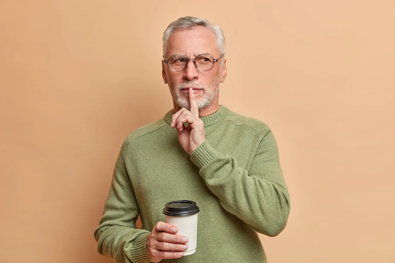 An older man in glasses holds a finger to his lips while holding coffee.