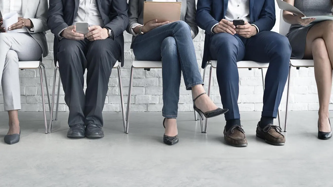 A row of people in business clothes wait in chairs with their phones.