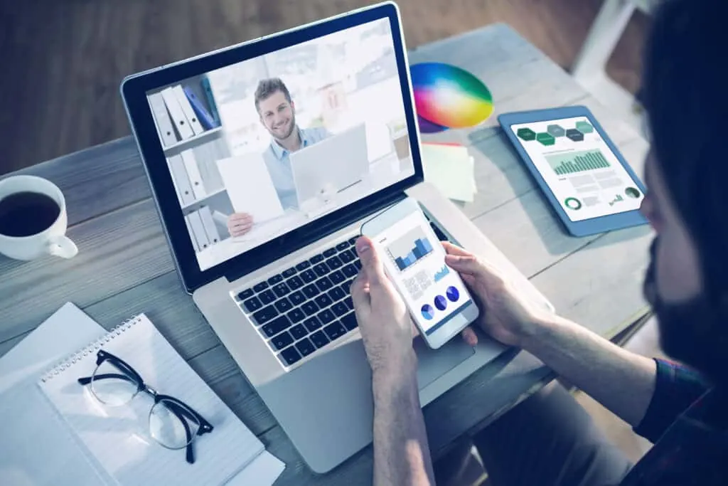A person holds a phone in front of a laptop showing a video call.