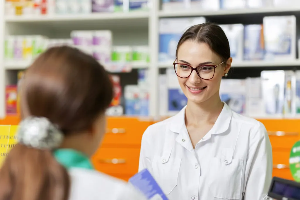A smiling pharmacist in a white coat helps a customer at a pharmacy counter.