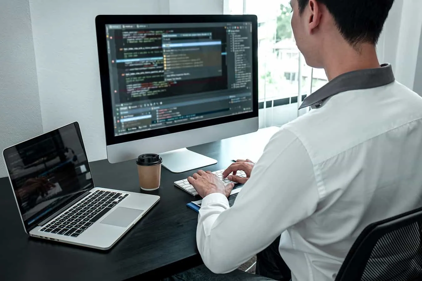 A programmer writes code on a desktop monitor beside an open laptop.