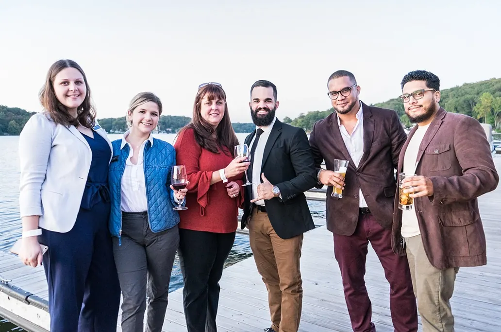 Six smartly dressed people pose together with drinks at a waterfront event.