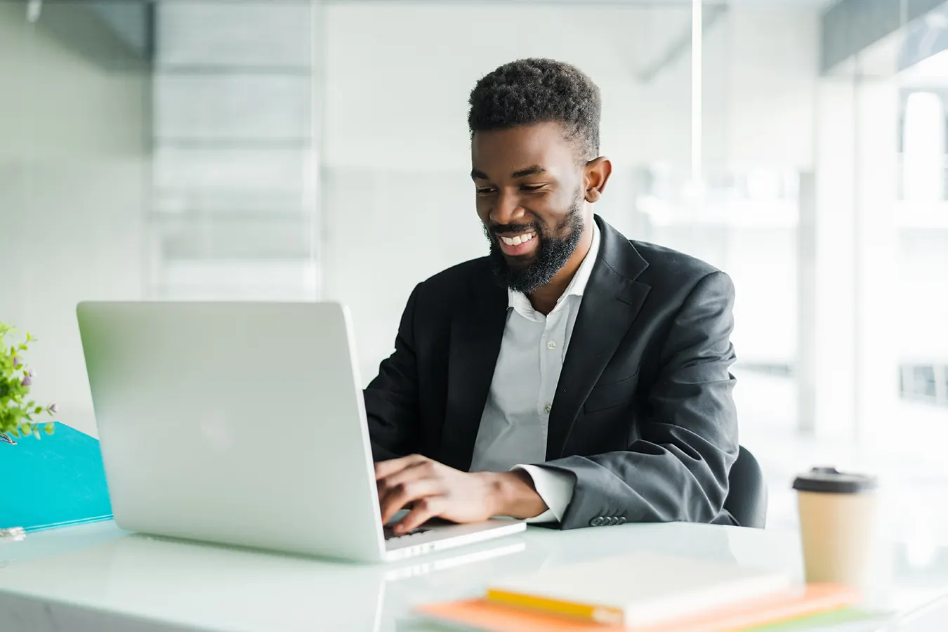 A smiling businessman in a suit types on a laptop at a white desk.