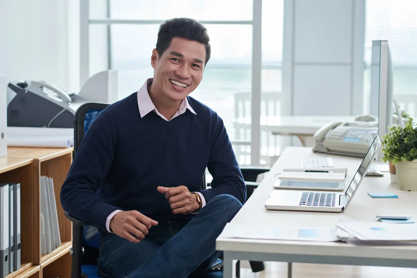 A smiling man in a navy sweater sits at an office desk.