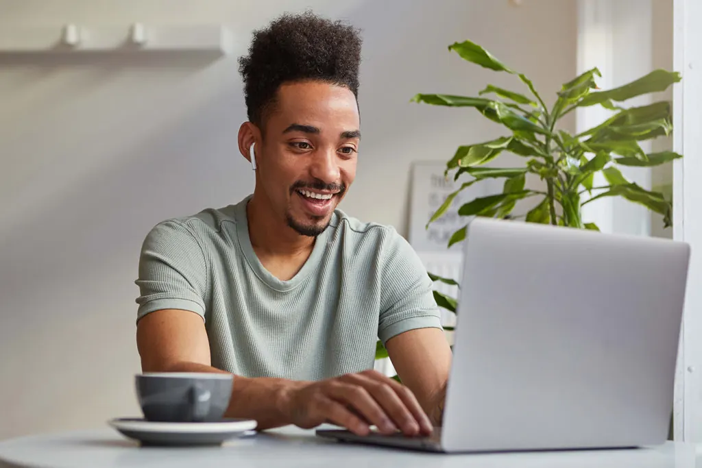 A smiling young man works on a laptop at a table with a plant.