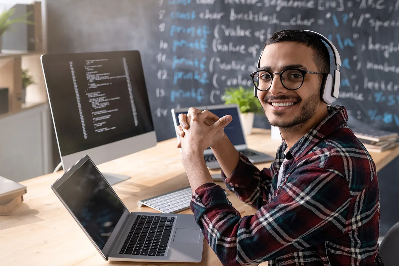 A smiling programmer with headphones sits at a desk with a laptop and code.