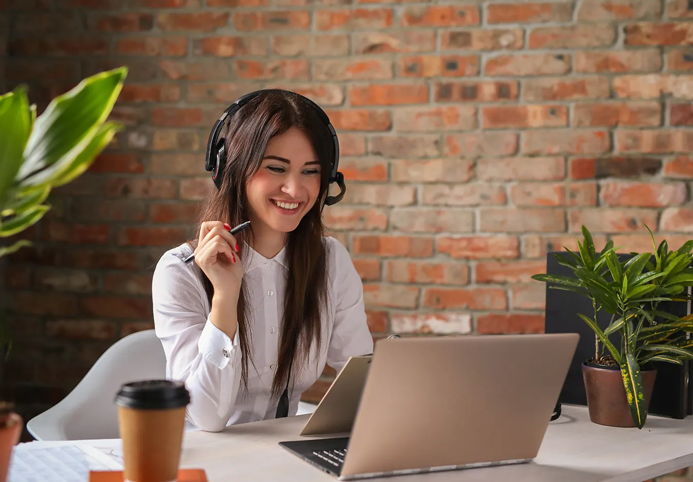 A smiling woman with a headset works at a laptop by a brick wall.