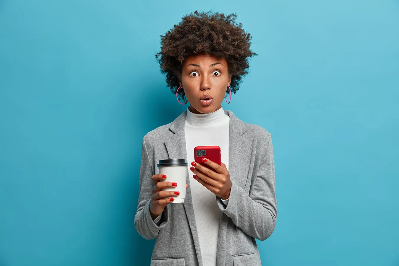 A surprised woman holds a phone and a coffee cup against a blue wall.