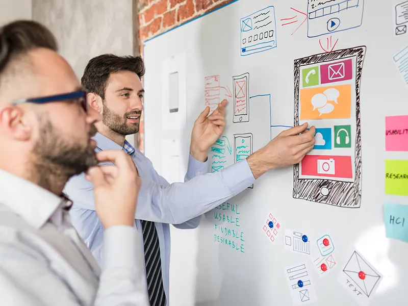 Two men plan an app design on a whiteboard covered in sketches.