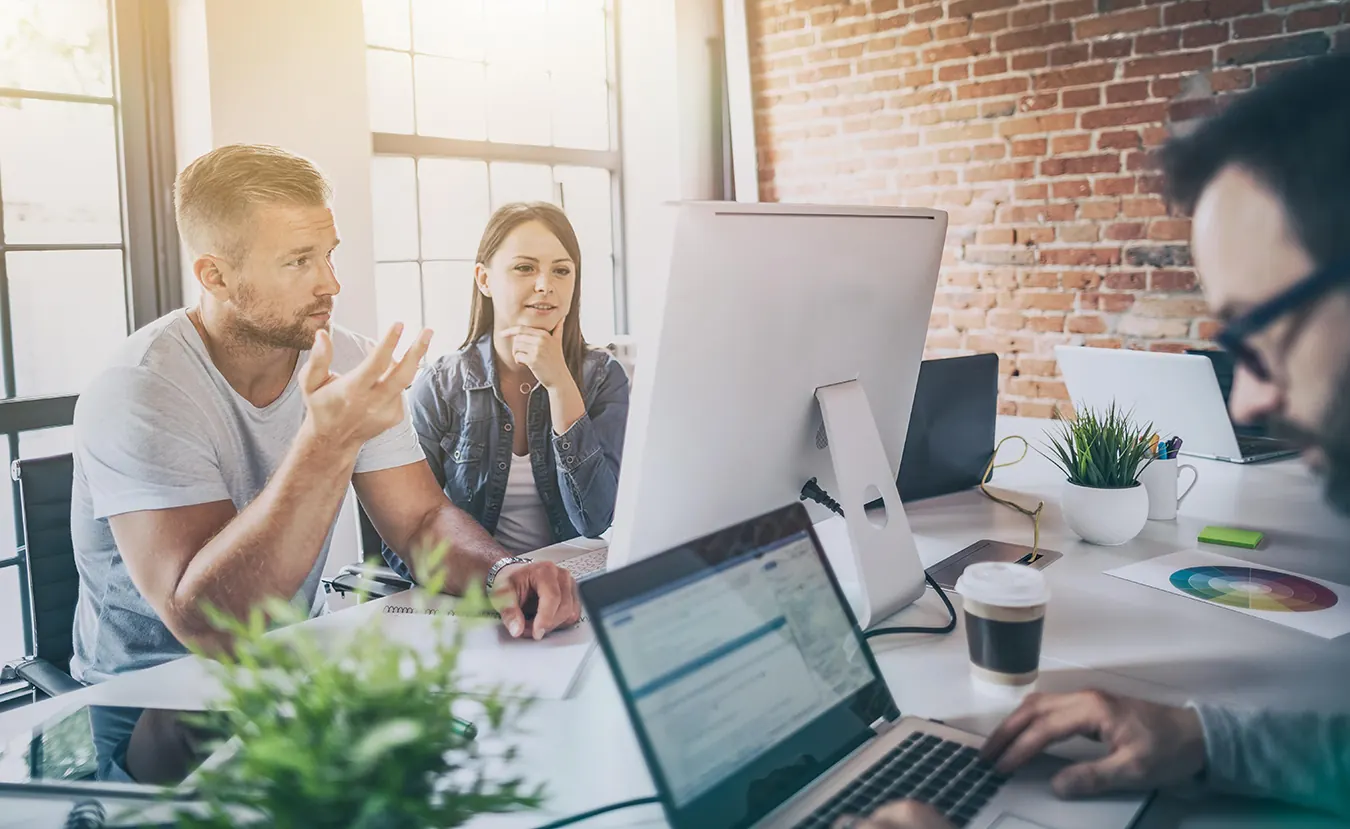 Three coworkers talk at a shared office desk with laptops and monitors.