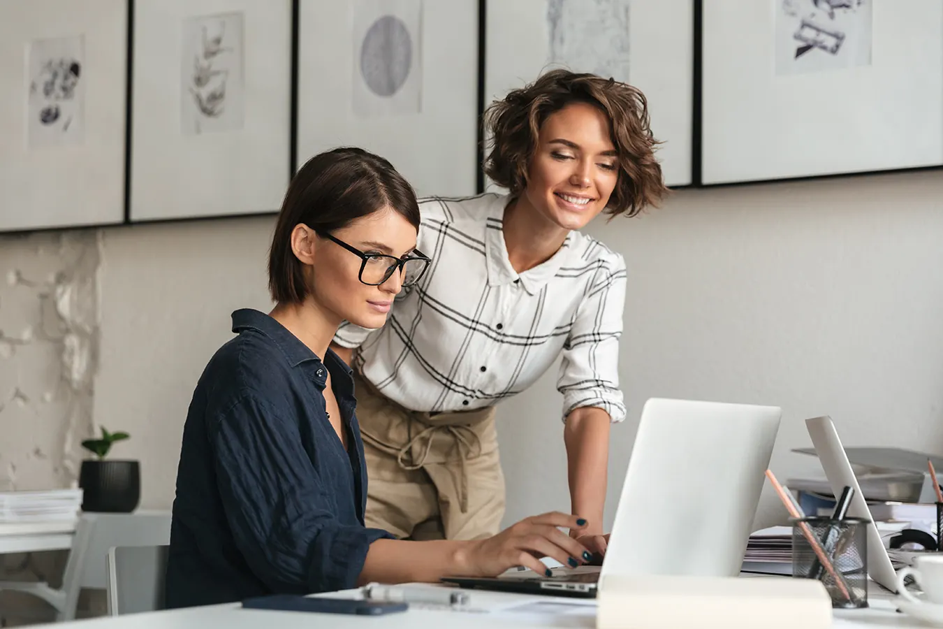 Two women discuss work together at a laptop in a bright office.