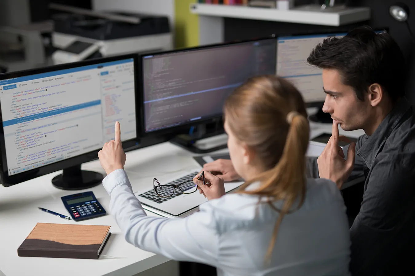 A woman points at code on a monitor while a man watches beside her.
