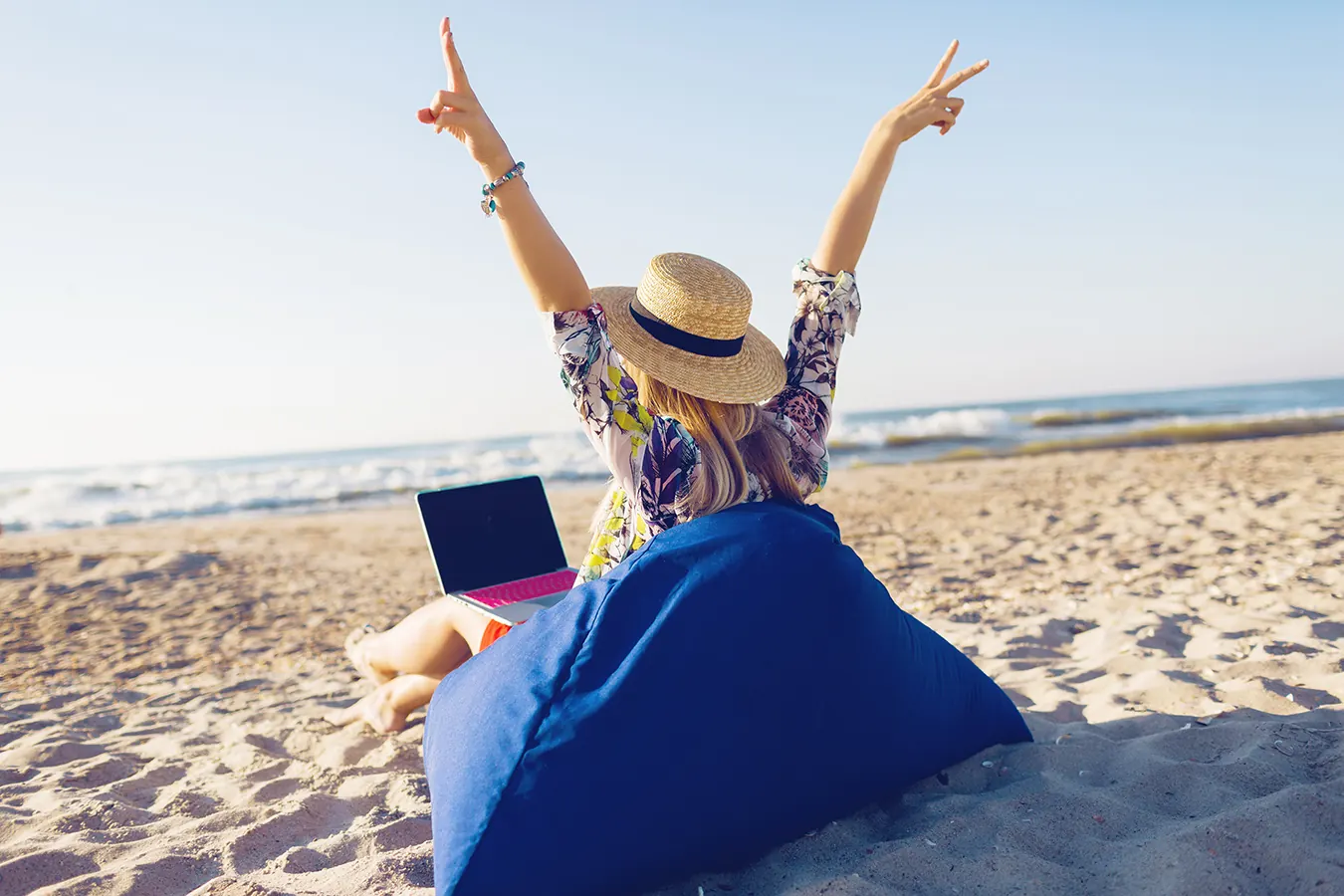 A woman in a sun hat raises her arms beside a laptop on sand.