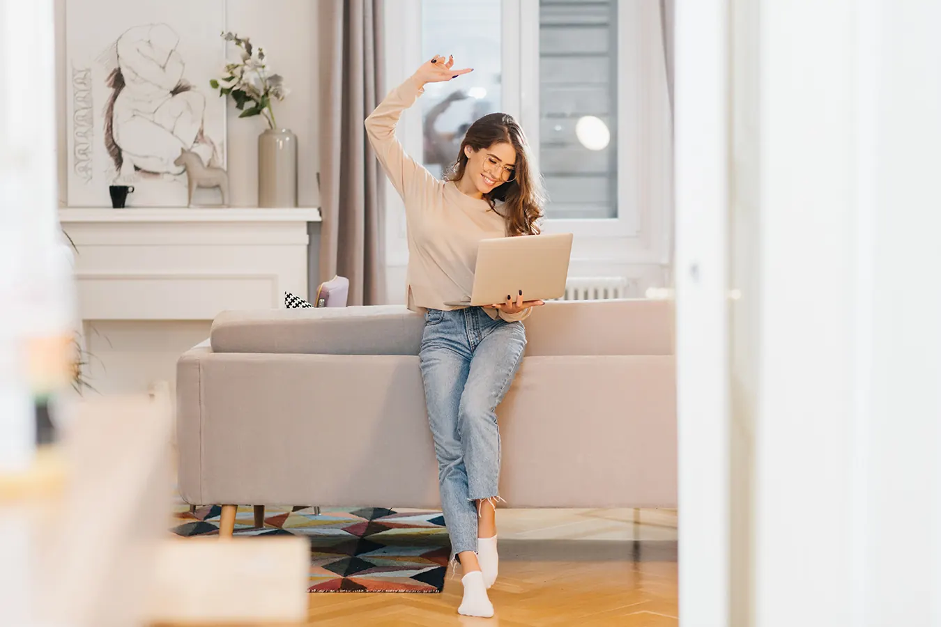 A happy woman dances while holding a laptop in a bright living room.
