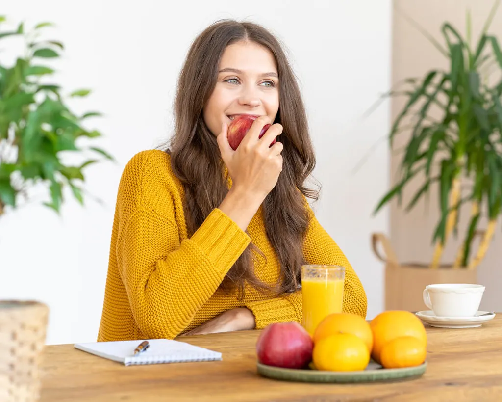 A smiling woman in a yellow sweater bites an apple beside fruit.