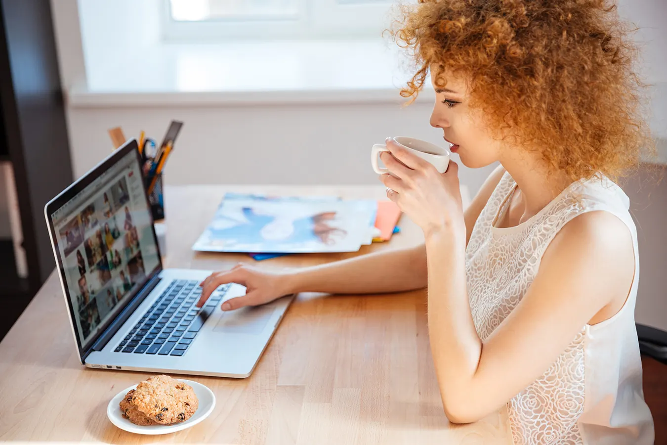 A woman with curly hair sips coffee while looking at her laptop.