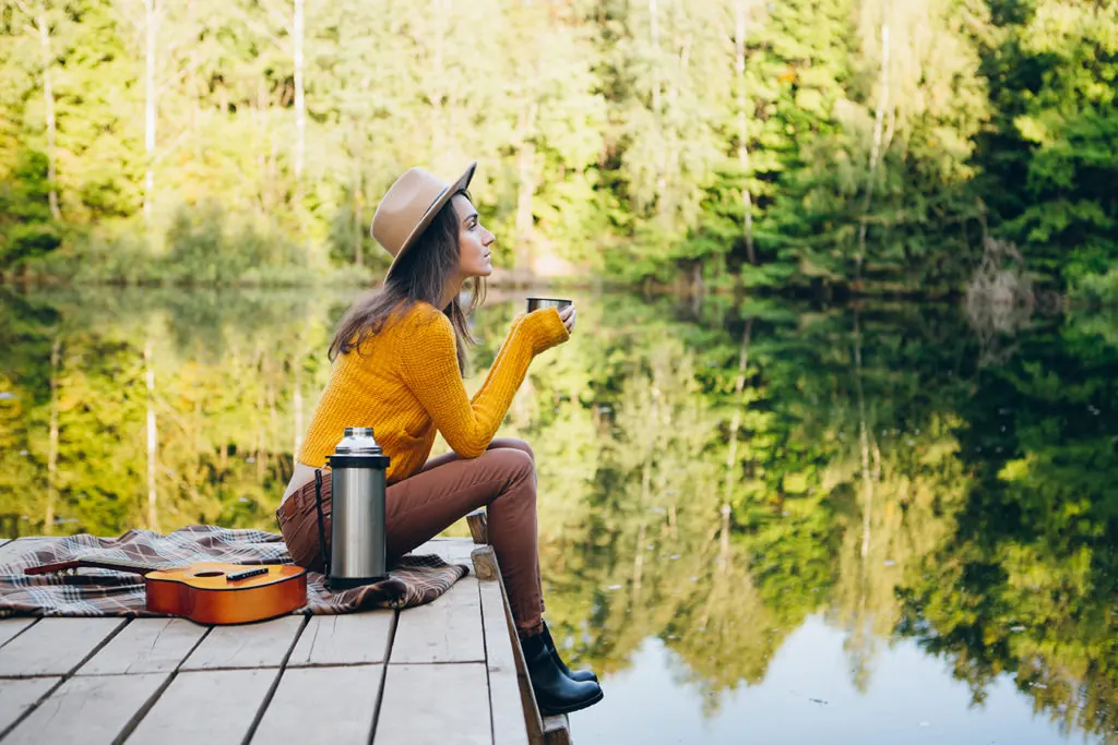 A woman in a yellow sweater sits on a dock beside a forest lake.