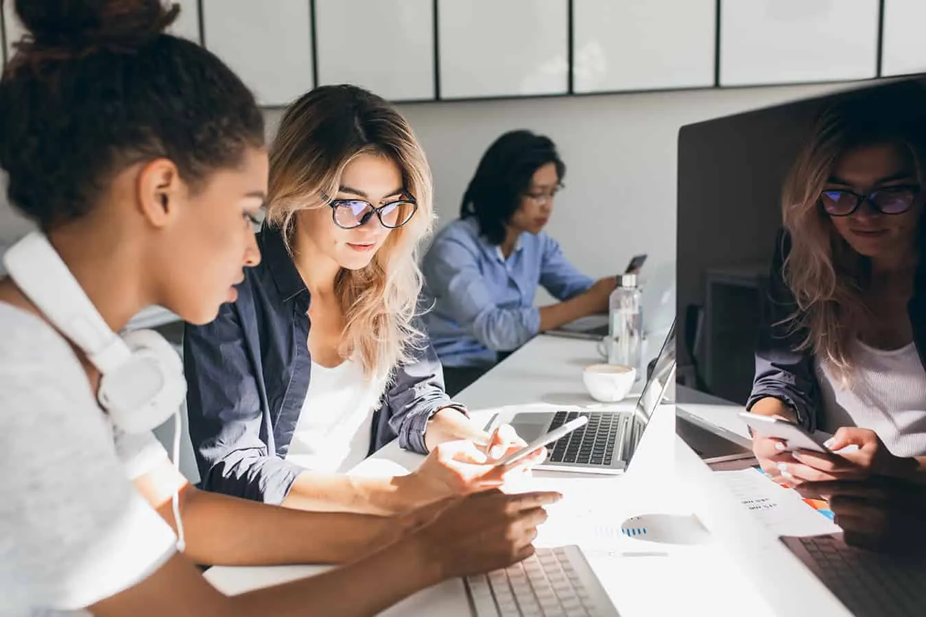 A group of women work on laptops and phones at an office table.