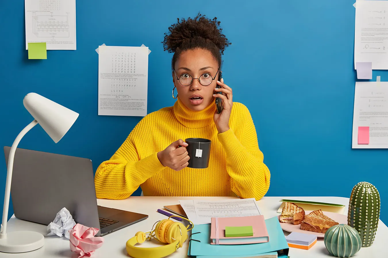 A worried woman in a yellow sweater talks on the phone at her desk.