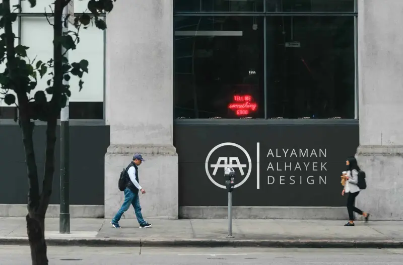 The Alyaman Alhayek Design logo on a storefront wall as people walk past.