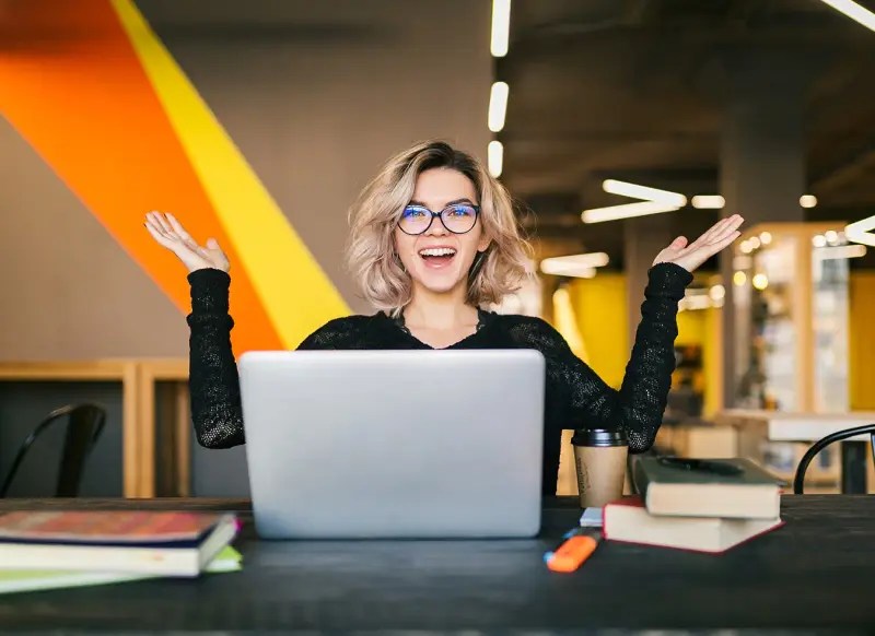An excited woman in glasses raises her hands behind a laptop in a cafe.