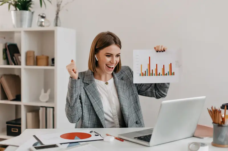 A happy businesswoman at her laptop holds up a printed growth chart.