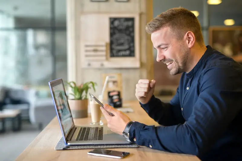 A happy man celebrates while looking at his phone beside a laptop.