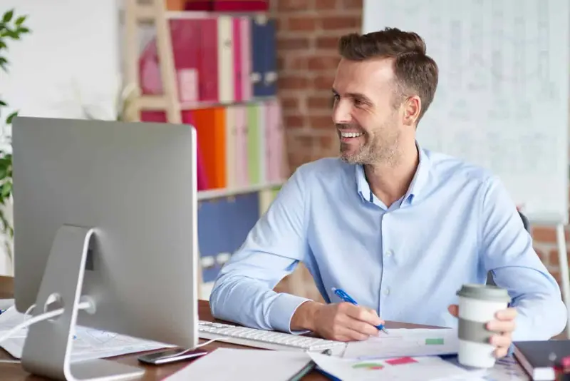 A smiling man works at a desktop computer in a bright office.