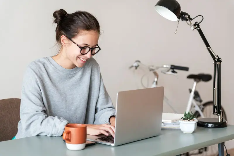 A smiling woman in glasses works on a laptop at a home desk.