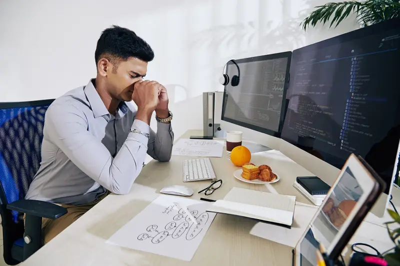 A stressed programmer rubs his eyes at a desk with code on two monitors.